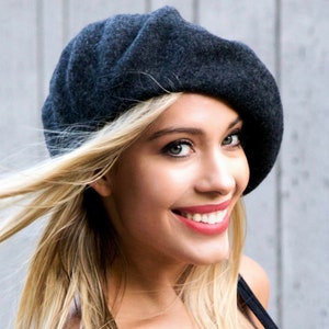 May include: A woman with long blonde hair is wearing a dark gray beret. She is smiling and looking to the right.