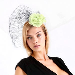 May include: A fascinator with a light green flower and black netting. The headpiece is set against a white background. The flower is a soft, textured material. The netting is a delicate, open weave.