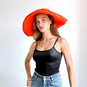 May include: A wide-brimmed, bright orange sun hat. The hat has a large, circular brim and a rounded crown. The hat is worn with a black tank top and blue jeans. The background is a plain white.