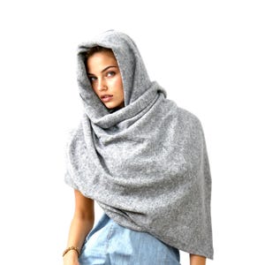 May include: A light gray hooded scarf, draped around the shoulders. The scarf is made of a soft, textured material. The hood is up, creating a cozy and stylish look. The scarf is a versatile accessory for cooler weather.