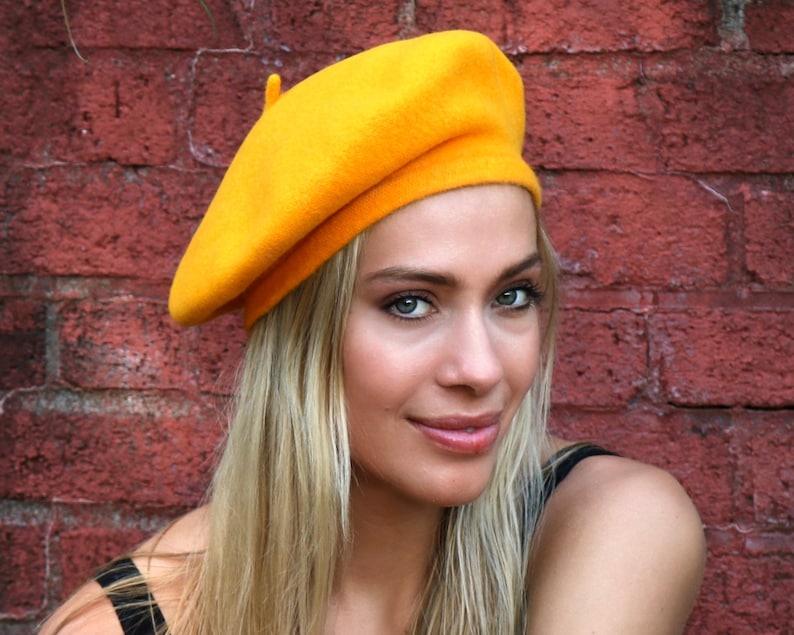 May include: A woman wearing a bright yellow beret. The beret is made of felt and has a classic style. The woman has long blonde hair and is wearing a black tank top.