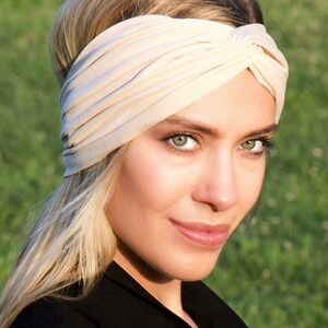 May include: A beige turban-style headband. The fabric is soft and gathered, creating a textured appearance. The headband is worn on the head. The background is a blurred green field.