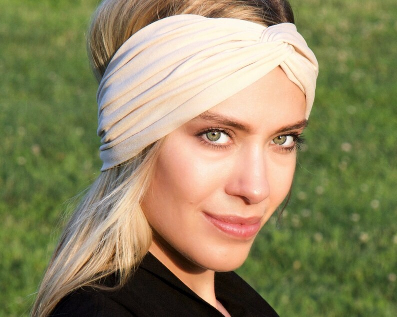May include: A beige turban-style headband. The fabric is soft and gathered, creating a textured appearance. The headband is worn on the head. The background is a blurred green field.