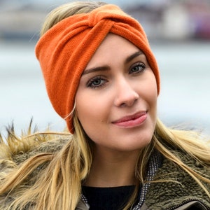 May include: A woman with long blonde hair wearing a bright orange knitted headband. The headband is tied in a knot on top of her head.
