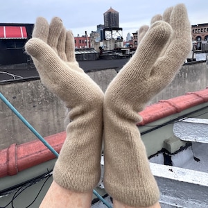 May include: A pair of beige knitted gloves with a ribbed texture. The gloves are held up in the air with the fingers spread out.