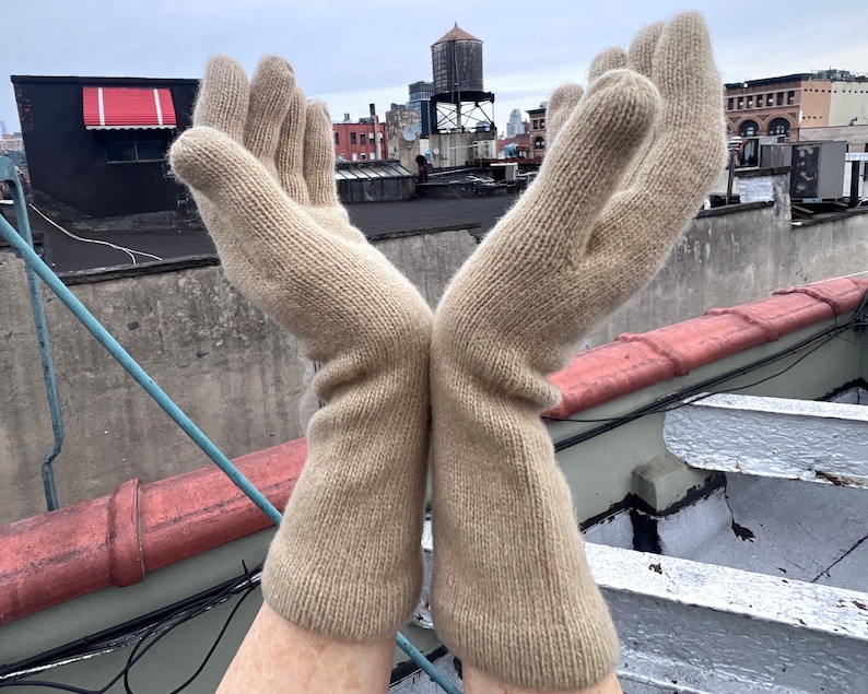 May include: A pair of beige knitted gloves with a ribbed texture. The gloves are held up in the air with the fingers spread out.