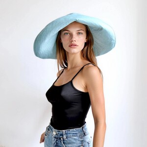 May include: A light blue, wide-brimmed sun hat. The hat has a large, floppy brim and a rounded crown. The model is wearing a black tank top and blue jeans. The background is a plain white wall.