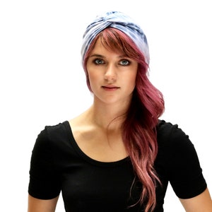 May include: A woman wearing a blue and white tie-dye turban and a black scoop-neck shirt. Her hair is a reddish-pink color and falls over her shoulder. The background is plain white.