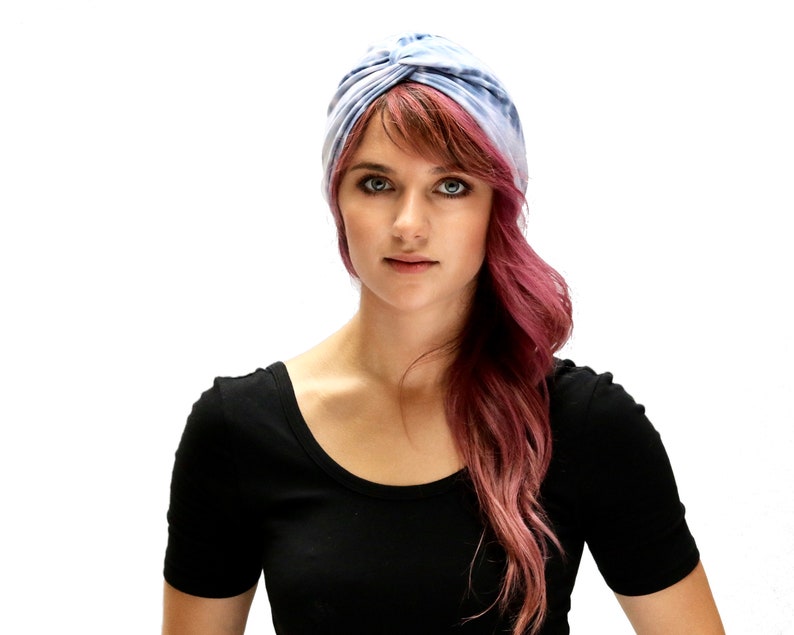 May include: A woman wearing a blue and white tie-dye turban and a black scoop-neck shirt. Her hair is a reddish-pink color and falls over her shoulder. The background is plain white.