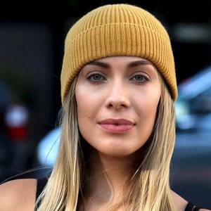 May include: A mustard yellow ribbed knit beanie. The hat is worn by a person with long blonde hair and fair skin. The background is blurred, with hints of a dark structure and a vehicle.