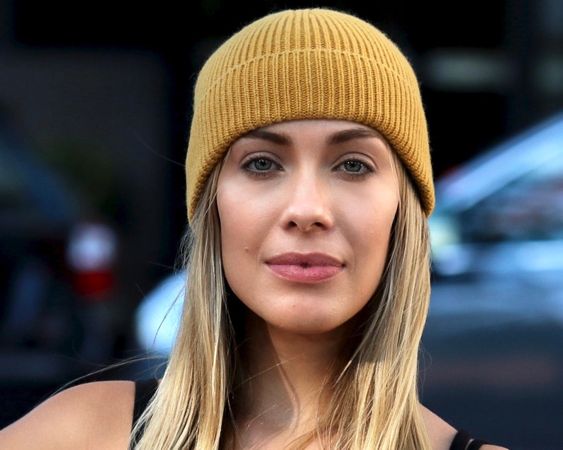 May include: A mustard yellow ribbed knit beanie. The hat is worn by a person with long blonde hair and fair skin. The background is blurred, with hints of a dark structure and a vehicle.
