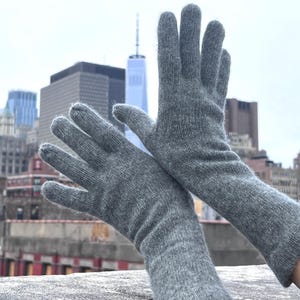 May include: A pair of long gray cashmere gloves with a ribbed texture. The gloves are shown against a city skyline background.