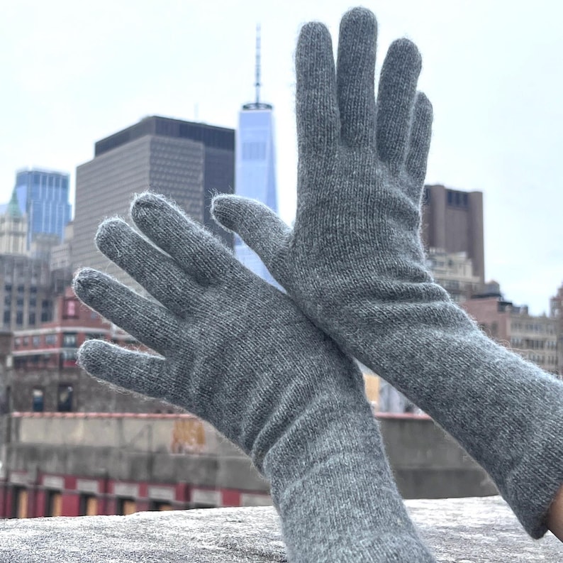 May include: A pair of long gray cashmere gloves with a ribbed texture. The gloves are shown against a city skyline background.