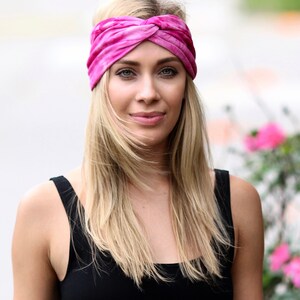 May include: A pink tie-dye headband is worn across the forehead. The headband is made of a soft fabric and has a twisted knot design. The person is wearing a black tank top.