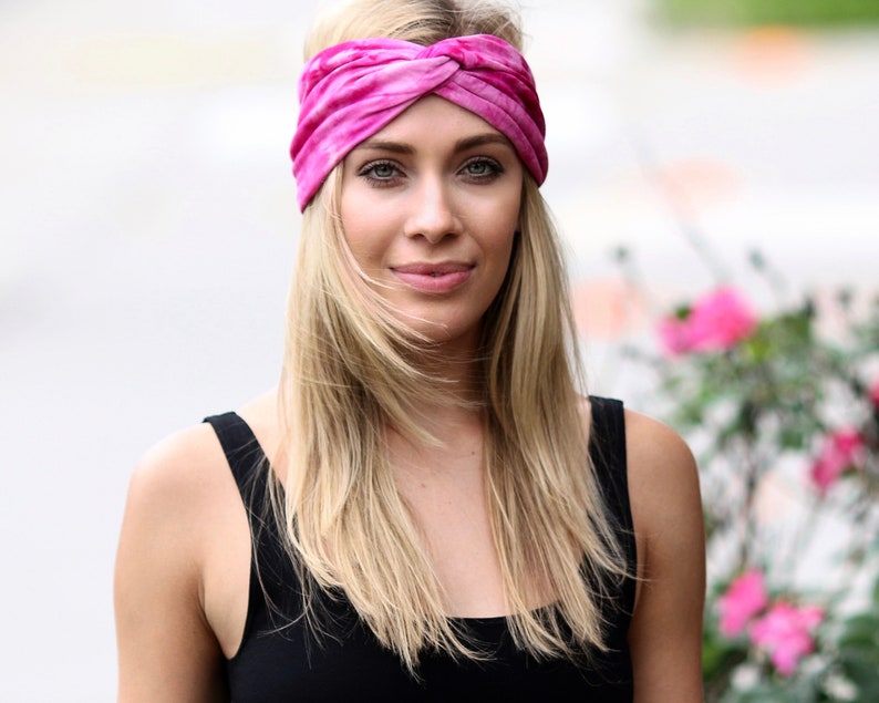 May include: A pink tie-dye headband is worn across the forehead. The headband is made of a soft fabric and has a twisted knot design. The person is wearing a black tank top.