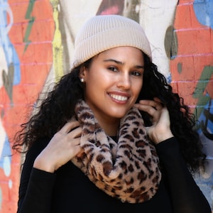 May include: A woman wearing a tan beanie and a faux fur leopard print infinity scarf. The woman is wearing a black long-sleeve shirt. The background is a colorful graffiti wall.