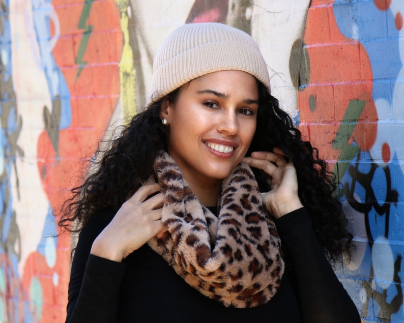 May include: A woman wearing a tan beanie and a faux fur leopard print infinity scarf. The woman is wearing a black long-sleeve shirt. The background is a colorful graffiti wall.
