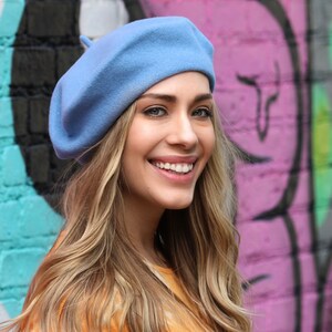 May include: A woman with long blonde hair is wearing a light blue beret. She is smiling and looking at the camera. The background is a pink and green graffiti wall.
