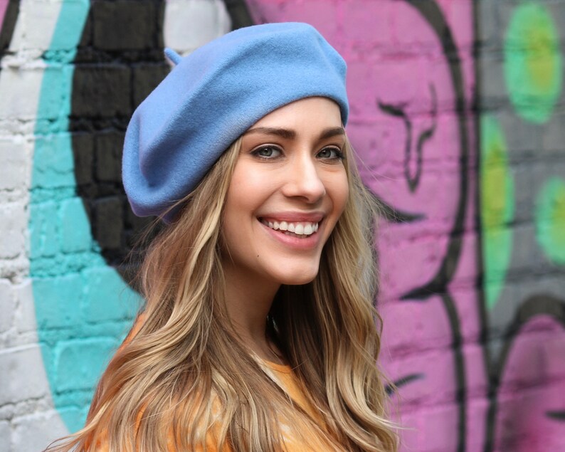 May include: A woman with long blonde hair is wearing a light blue beret. She is smiling and looking at the camera. The background is a pink and green graffiti wall.