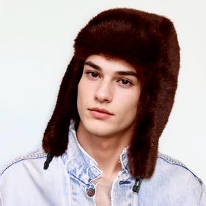 May include: A brown faux fur Ushanka hat with ear flaps. The hat is a dark shade of brown and has a soft, textured appearance. The hat is worn with a light blue denim jacket.