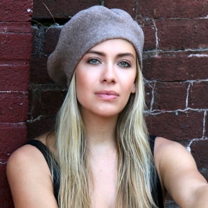 May include: A woman with long blonde hair is wearing a light brown beret. She is standing in front of a brick wall.