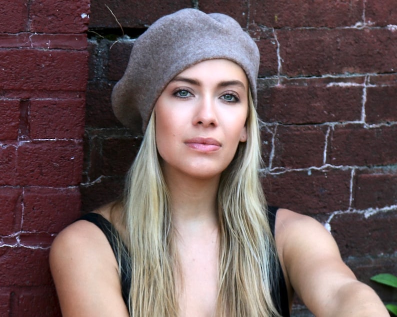 May include: A woman with long blonde hair is wearing a light brown beret. She is standing in front of a brick wall.