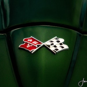 May include: A close-up of a silver Corvette emblem with a red and white checkered flag design on a green car hood.