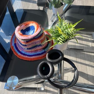 May include: A colorful, crocheted hat with a wide brim, featuring a vibrant mix of orange, blue, pink, and purple yarns. Black over-ear headphones are also visible. The items are on a glass table with plants in the background.