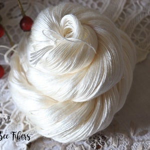 Puede incluir: Una madeja de hilo de seda blanco, enrollada en forma de bola, sobre un fondo de encaje blanco. El hilo es suave y delicado, y la madeja tiene un di&aacute;metro de unos 25 cm.