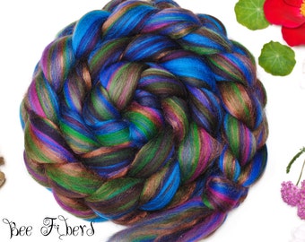 Puede incluir: Una trenza de fibra teñida a mano en tonos vibrantes de azul, verde, morado y marrón. A la izquierda hay un huso de madera con un envoltorio de fibra marrón. El texto "Beesy Bee Fibers" está en la parte inferior izquierda. Flores rojas y rosas y hojas verdes.
