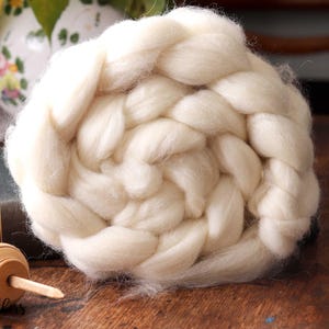 May include: A large, off-white braid of wool roving, tightly wound in a spiral. A wooden spindle and a vintage book are in the foreground. The background includes a vase with flowers. The text "Beesy Bee Fibers" is visible in the lower left corner.