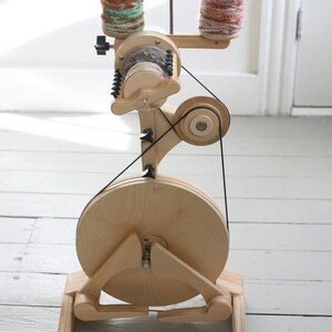 May include: A wooden spinning wheel with a large wooden wheel and a smaller wheel with a belt connecting them. Two spools of yarn are on the top of the spinning wheel. The yarn is multicolored.