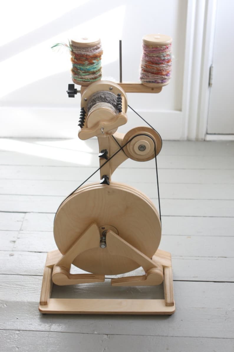 May include: A wooden spinning wheel with a large wooden wheel and a smaller wheel with a belt connecting them. Two spools of yarn are on the top of the spinning wheel. The yarn is multicolored.