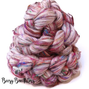 May include: A large, twisted skein of colorful yarn. The yarn is a blend of white, pink, purple, and blue fibers, with strands of silver metallic thread. The text "Beesy Bee Fibers" is printed in black script at the bottom.