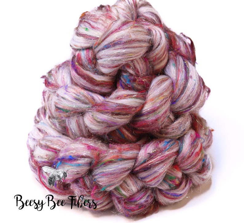 May include: A large, twisted skein of colorful yarn. The yarn is a blend of white, pink, purple, and blue fibers, with strands of silver metallic thread. The text "Beesy Bee Fibers" is printed in black script at the bottom.