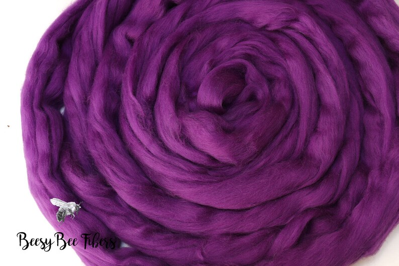 May include: A close-up of a deep purple wool roving, a type of fiber used for spinning yarn or felting. The roving is arranged in a spiral shape, highlighting its soft and fluffy texture.