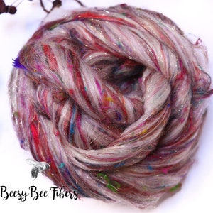 May include: A close-up of a skein of colorful yarn. The yarn is a mix of light gray, red, and purple with flecks of blue, green, and gold. The yarn has a sparkly, shiny appearance. The text "Beesy Bee Fibers" is in the bottom left corner.