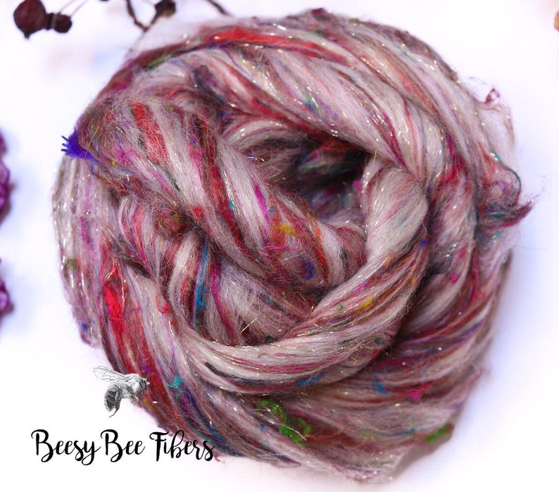 May include: A close-up of a skein of colorful yarn. The yarn is a mix of light gray, red, and purple with flecks of blue, green, and gold. The yarn has a sparkly, shiny appearance. The text "Beesy Bee Fibers" is in the bottom left corner.