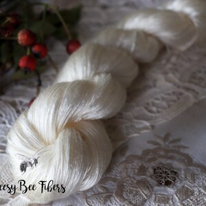 Puede incluir: Una madeja de hilo de seda blanco, retorcido y en bucle, con el texto "Beesly Bee Fibers" en letras negras.