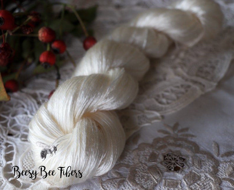 Puede incluir: Una madeja de hilo de seda blanco, retorcido y en bucle, con el texto "Beesly Bee Fibers" en letras negras.