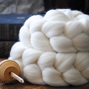 May include: A white, fluffy braid of wool fiber, ready for spinning, sits next to a wooden spinning wheel.
