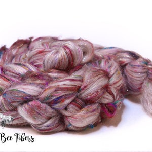 May include: A braided skein of colorful yarn, featuring a blend of pink, red, blue, and white fibers with a shimmering, iridescent quality. The yarn is arranged in a spiral, with the text "Beesy Bee Fibers" in the lower left corner.