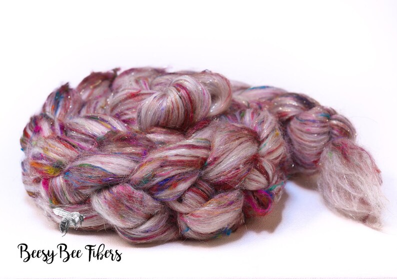 May include: A braided skein of colorful yarn, featuring a blend of pink, red, blue, and white fibers with a shimmering, iridescent quality. The yarn is arranged in a spiral, with the text "Beesy Bee Fibers" in the lower left corner.
