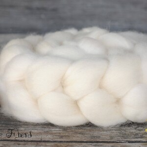 Puede incluir: Un gran paquete trenzado de mechas de lana blanco crema. Las fibras suaves y esponjosas son ideales para hilar o afieltrar. La imagen tambi&eacute;n incluye un objeto decorativo con pintura roja y blanca y el texto "Beesy Bee Fibers".