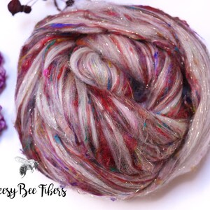 May include: A close-up of a ball of colorful yarn with a mix of red, pink, blue, and silver threads. The yarn is a blend of fibers, with a soft, textured appearance. The logo "Beesy Bee Fibers" is in the bottom left corner.