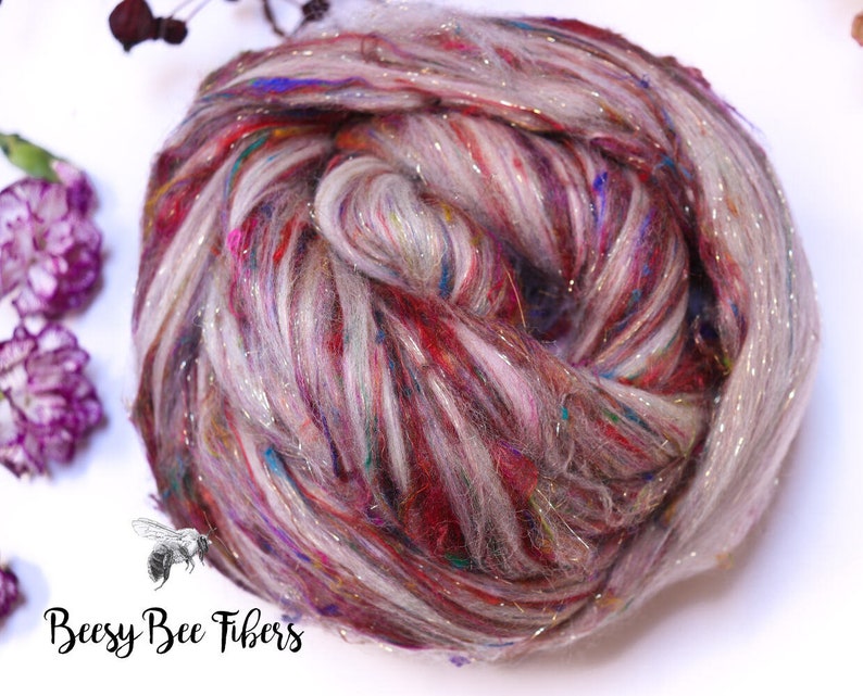 May include: A close-up of a ball of colorful yarn with a mix of red, pink, blue, and silver threads. The yarn is a blend of fibers, with a soft, textured appearance. The logo "Beesy Bee Fibers" is in the bottom left corner.