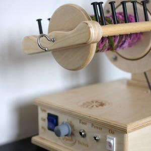 May include: A wooden spinning wheel with a pink and green yarn skein on the spindle. The wheel has a foot pedal and a speed control knob. The power switch is labeled "On" and "Off".