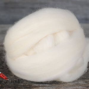 Puede incluir: Una gran y esponjosa nube de mecha de lana sin te&ntilde;ir, de color blanquecino, se encuentra junto a un huso de madera con detalles pintados en rojo y blanco. La lana es suave y parece lista para hilar o afieltrar. La imagen est&aacute; sobre un fondo de madera r&uacute;stica.