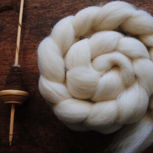 Puede incluir: Una lana blanca en roving est&aacute; enrollada en espiral, lista para hilar. Tambi&eacute;n se muestra una rueca de madera con un huso marr&oacute;n.