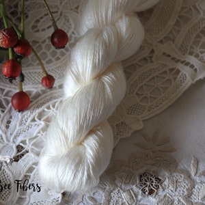 Puede incluir: Un ovillo de hilo de seda blanco, presentado sobre un encaje blanco con el texto "Beesly Bee Fibers" en la esquina inferior izquierda.